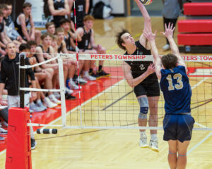 High school boys' volleyball match: player spikes ball over the net, while teammates and spectators watch intently.