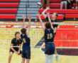 High school volleyball players in action, one spiking the ball over the net while two opponents jump to block.