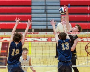 A high school volleyball match with players jumping at the net to block a spike. Excitement fills the gym.