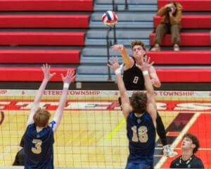 High school volleyball match with player #8 spiking the ball over the net as defenders jump to block.