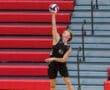 Male volleyball player serves the ball while leaping against a backdrop of red bleachers.