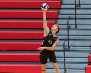 Male volleyball player serves the ball while leaping against a backdrop of red bleachers.