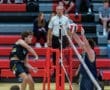 High school volleyball players competing at the net, with spectators in the background watching the intense match.