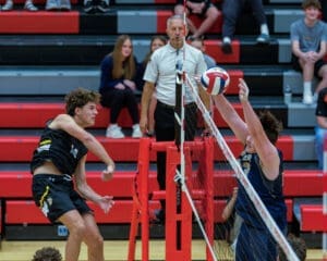 High school volleyball players competing at the net, with spectators in the background watching the intense match.