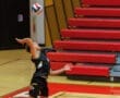 A volleyball player serves the ball high in a gym with red bleachers in the background, showcasing athletic focus and skill.