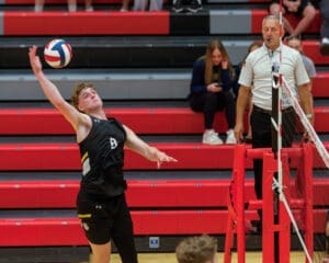 High school volleyball player serves the ball during a match, with the referee and spectators in the background.