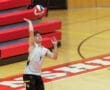 Young male volleyball player serving the ball during a match on a gym court with red bleachers in the background.