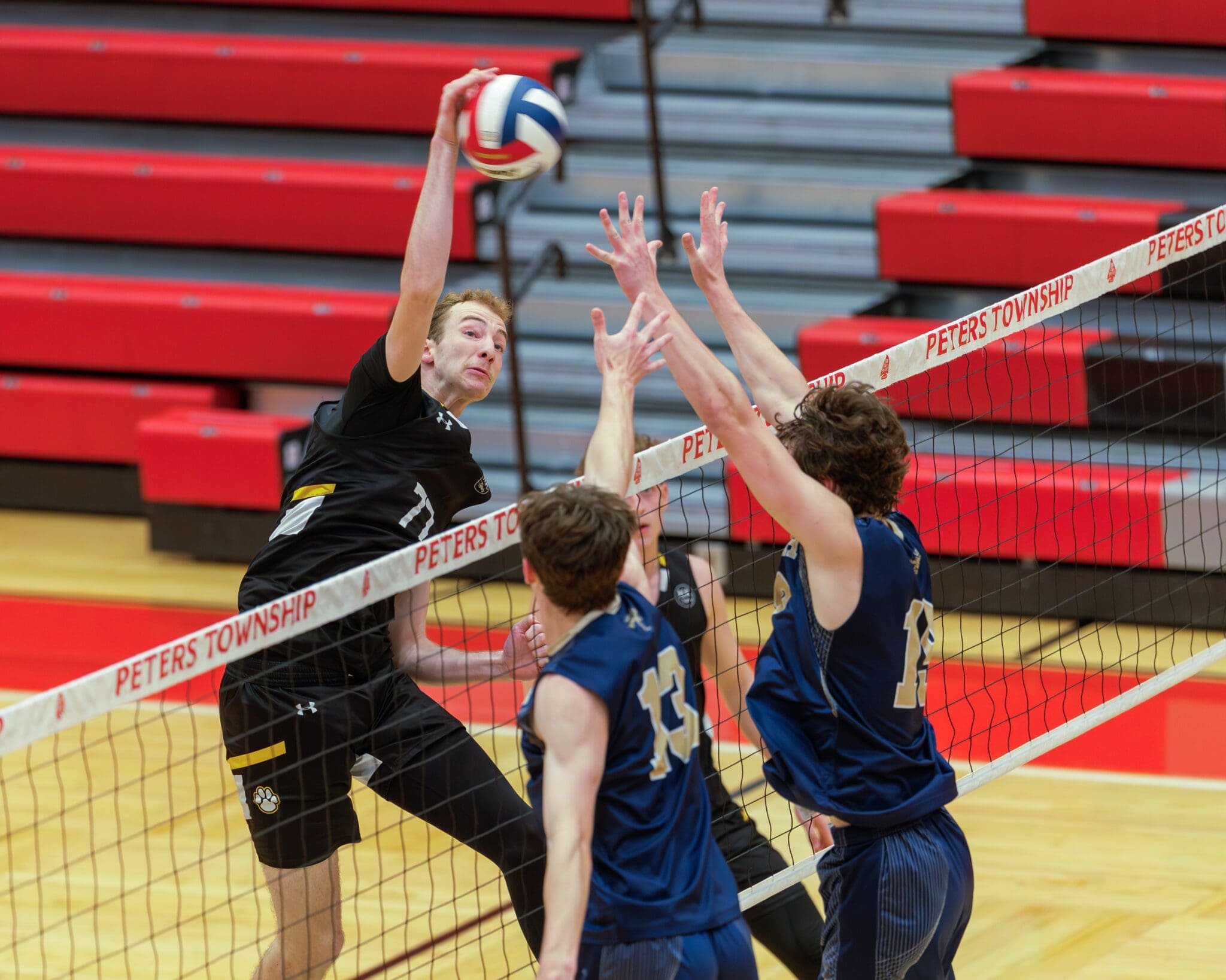High school volleyball match action, with a player spiking the ball over opposing defenders at the net.