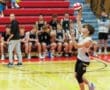 A young male volleyball player serves the ball during a match, with teammates watching from the sidelines.