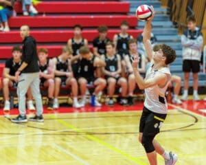 A young male volleyball player serves the ball during a match, with teammates watching from the sidelines.