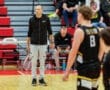 Basketball coach observing players during a game, with spectators in the background on a hardwood court.