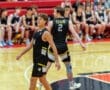 Two basketball players in black jerseys walking on the court, with teammates seated in the background.
