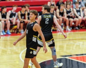 Two basketball players in black jerseys walking on the court, with teammates seated in the background.