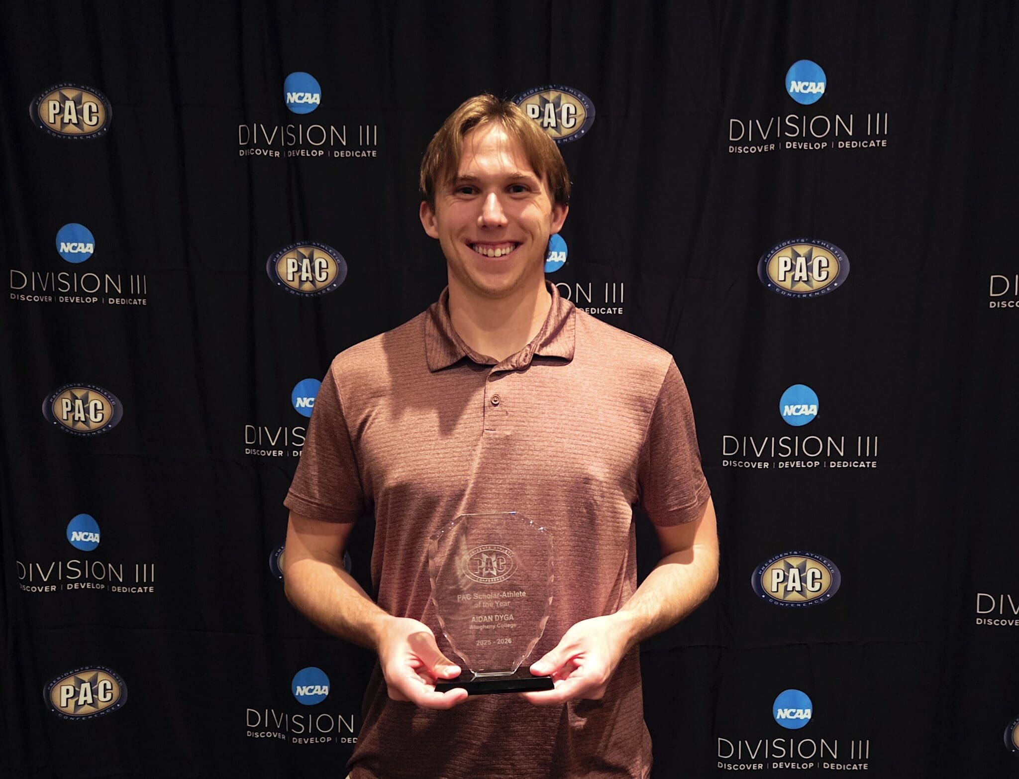 Aidan Dyga proudly holding the PAC Scholar Athlete of the Year award in front of a Division III backdrop.