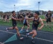 Two runners in a relay race at North Allegheny High School pass a baton as teammates cheer in the background on a track field.
