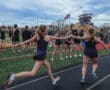 Track team members participate in a baton exchange during a relay race at North Allegheny High School, under a cloudy sky.