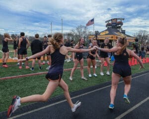Track team members participate in a baton exchange during a relay race at North Allegheny High School, under a cloudy sky.
