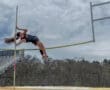 Female pole vaulter soaring over the bar at a track event, showcasing athleticism against a cloudy backdrop.