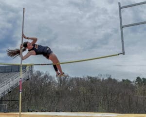 Female pole vaulter soaring over the bar at a track event, showcasing athleticism against a cloudy backdrop.