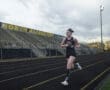 Young female athlete running on a track at North Allegheny, with stadium seating and cloudy skies in the background.