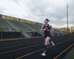 Young female athlete running on a track at North Allegheny, with stadium seating and cloudy skies in the background.