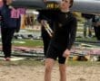 Young male rower carrying a boat on his shoulder at a rowing event, surrounded by equipment and spectators.