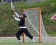 Goalkeeper in action during a lacrosse game, reaching for a high shot on a grassy field.