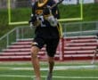 Young lacrosse player in a black jersey running on a turf field, prepared to make a play with his stick.