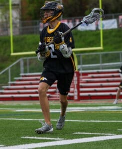 Young lacrosse player in a black jersey running on a turf field, prepared to make a play with his stick.
