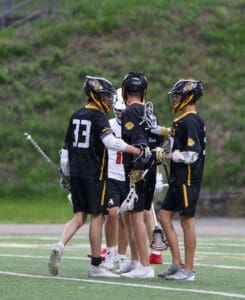 Boys' lacrosse team celebrates a play in the rain during a game on a green turf field.