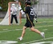 Lacrosse player in black jersey shoots on goal during a rainy game, with goalie preparing to block the shot.