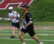 Lacrosse player number 16 runs with the ball on a field, while an opponent pursues in the background.