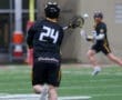 Lacrosse player in black jersey passes the ball on a wet field, with another player in the background ready to receive.