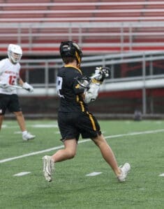 Lacrosse player in black jersey running on field during a rainy game, teammate in white jersey nearby.