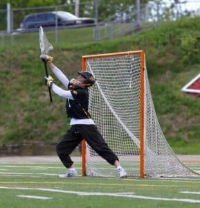 Goalkeeper in action during a lacrosse game, reaching for a high shot on a grassy field.