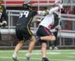 Lacrosse players compete on the field; one player is defending while another attempts to score in rainy conditions.