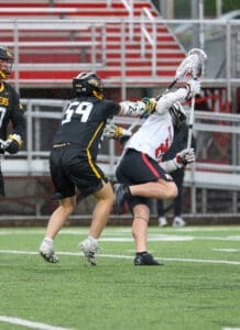 Lacrosse players compete on the field; one player is defending while another attempts to score in rainy conditions.