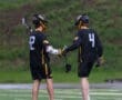 Two lacrosse players in black jerseys engage in a moment of camaraderie on the field during rainy weather.