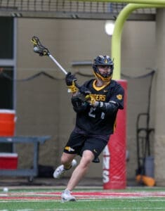 Lacrosse player in black jersey passes the ball during a game on a turf field, showcasing athleticism and teamwork.