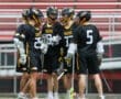 Lacrosse team huddled together in black and yellow uniforms, discussing strategy on the field.