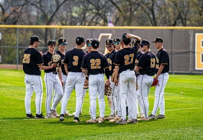 A group of young baseball players in matching black and gold uniforms huddle together on a sunny field.