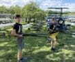 Two young rowers carrying oars in a boathouse area with a clear sky and lush greenery in the background.