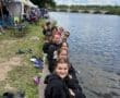 Group of smiling teens in black hoodies sitting by a river, enjoying the outdoors at a scenic event with tents nearby.
