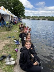 Group of smiling teens in black hoodies sitting by a river, enjoying the outdoors at a scenic event with tents nearby.