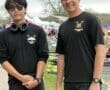 Two young rowers in black shirts pose together at a rowing event, with boats and tents in the background.