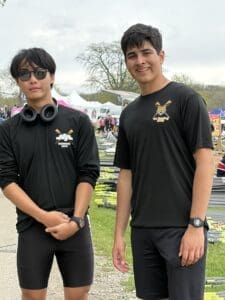 Two young rowers in black shirts pose together at a rowing event, with boats and tents in the background.