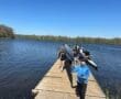 Group of people carrying kayaks on a sunny day at a lakeside dock, ready for an outdoor adventure.