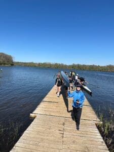 Group of people carrying kayaks on a sunny day at a lakeside dock, ready for an outdoor adventure.