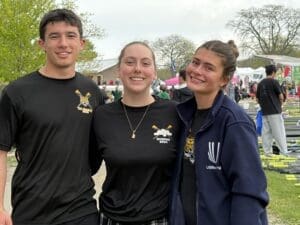Three young rowers smile together at a rowing event, showcasing camaraderie and team spirit in their athletic gear.