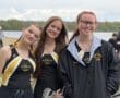 Three young female rowers pose for a photo by a lake, wearing matching NA Crew uniforms, smiling and cheerfully decorated.